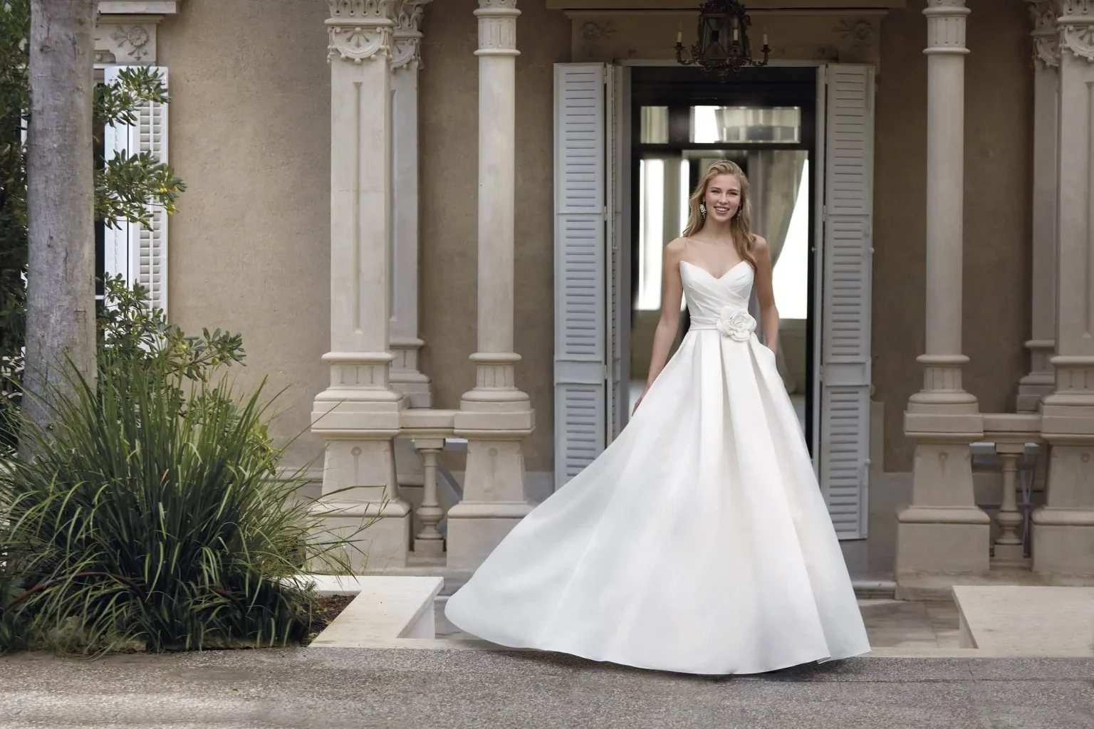 Extravagantes Brautkleid mit schmalem Trägerlos-Oberteil und voluminösem Rüschenrock auf antikem Sofa in der Natur, Modell ADYNIC – DIVAS Hochzeitshaus Singen